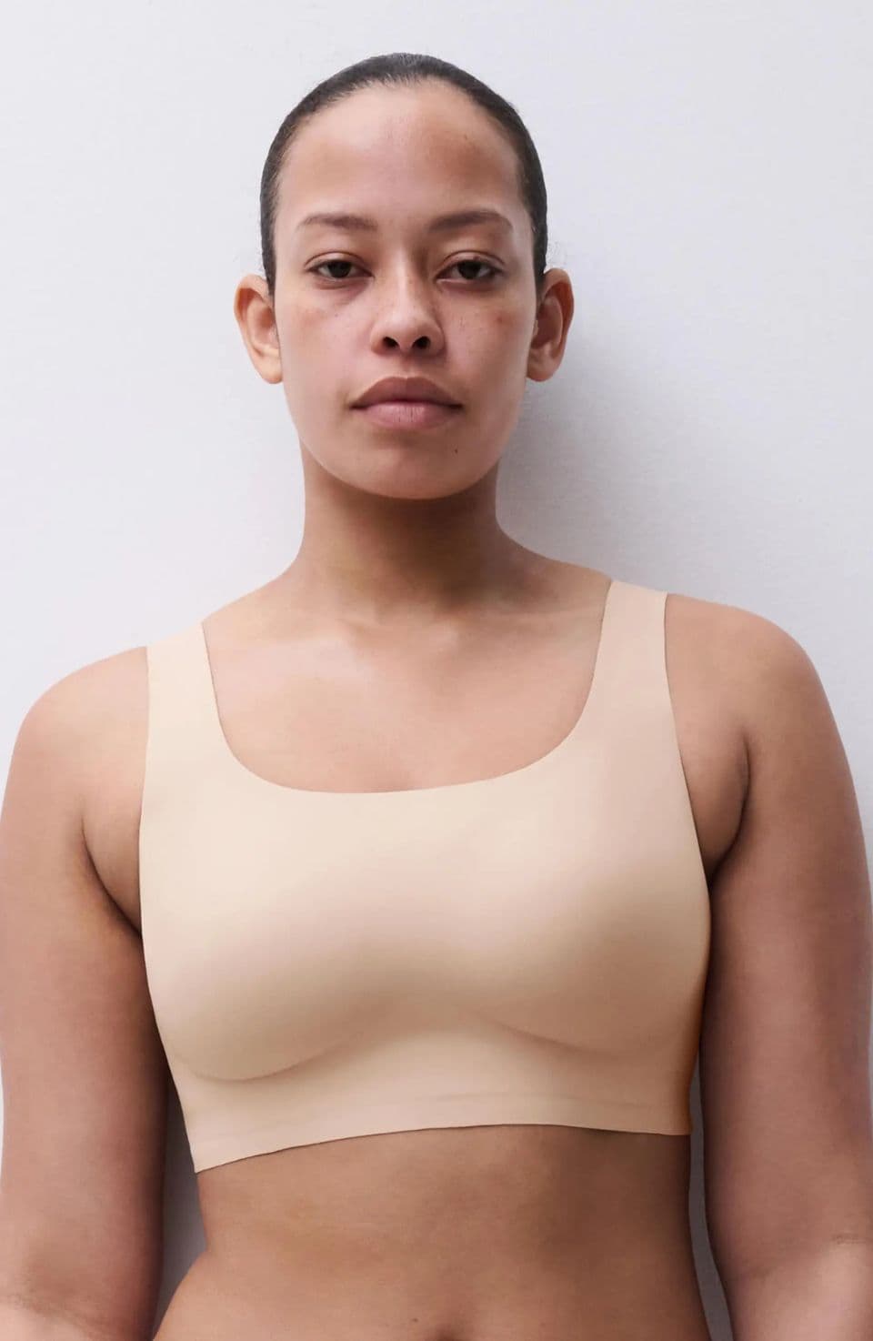 Person wearing a beige, seamless sports bra stands against a plain white background, looking directly at the camera.