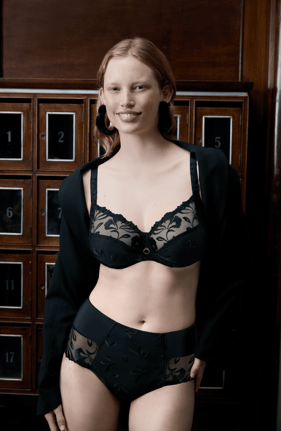 Smiling woman wearing a black lace lingerie set, standing in front of vintage wooden lockers.