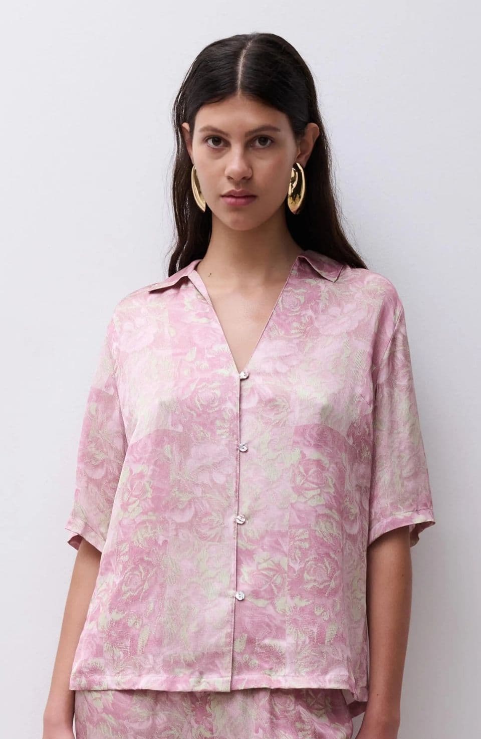 Woman wearing a pink patterned button-up shirt with short sleeves and gold hoop earrings, standing against a plain white background.