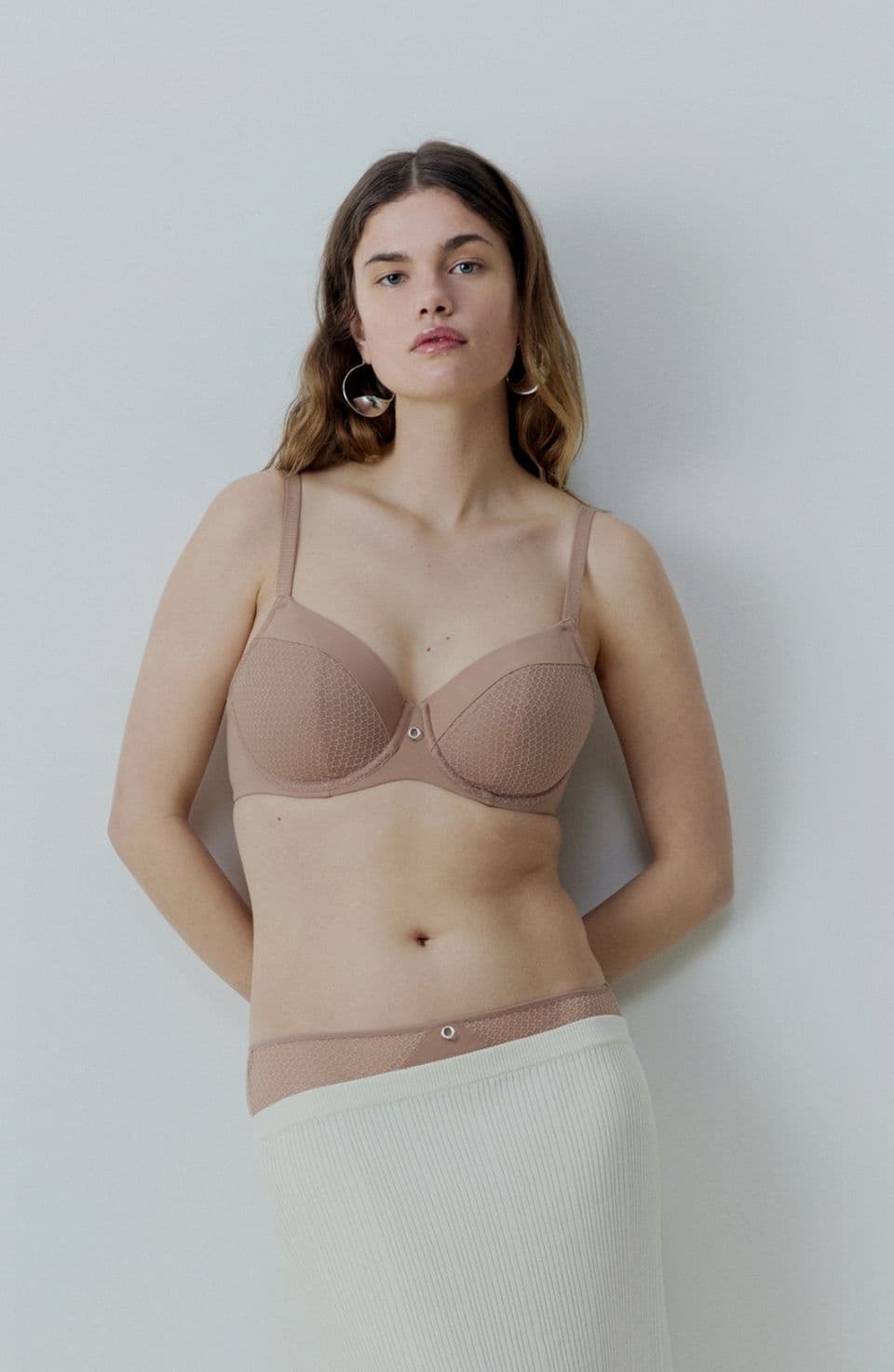 Woman wearing beige lingerie and a pleated skirt stands against a plain white background, looking confidently at the camera.