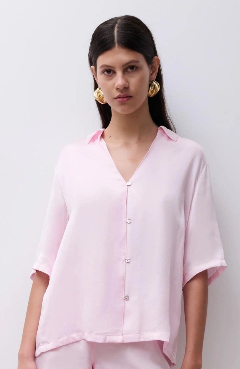 Woman wearing a light pink, short-sleeved button-up shirt with gold statement earrings, standing against a plain background.