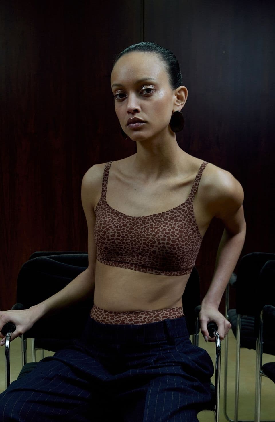 Person seated, wearing a brown leopard-print top and dark pants, against a dark wood panel background, looking confidently at the camera.