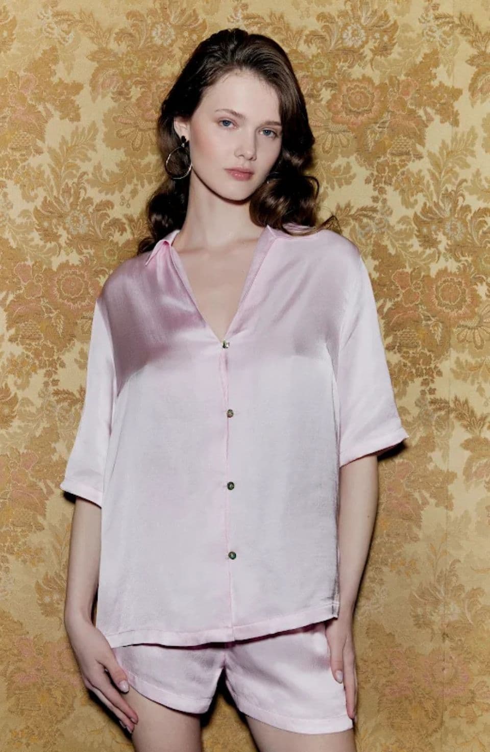 Woman in pink satin pajamas stands against a gold floral-patterned wallpaper, wearing hoop earrings and with long, wavy hair.