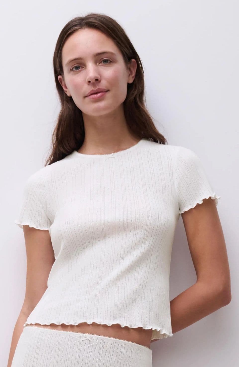 Person wearing a white, ribbed, short-sleeve top, smiling slightly, with long brown hair, standing against a plain light background.