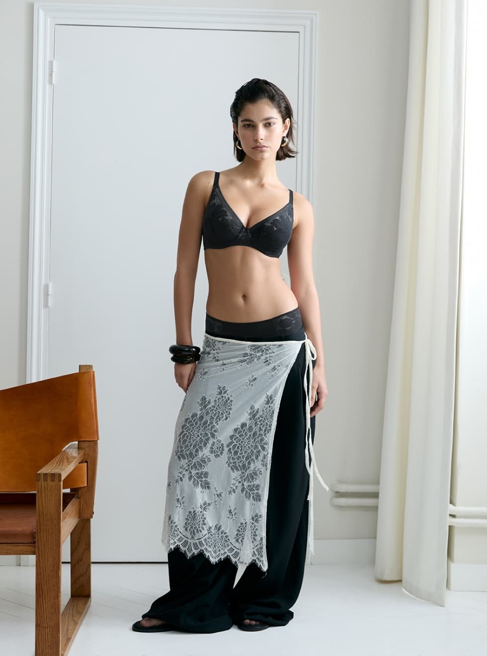 Woman in black bralette and pants with a lace overlay, standing near a wooden chair in a minimalist room.