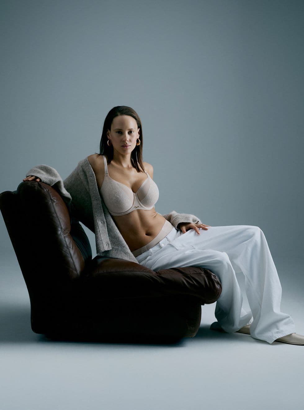 A woman in a beige bra, white pants, and a gray cardigan poses confidently seated on a brown leather chair in a softly lit room.