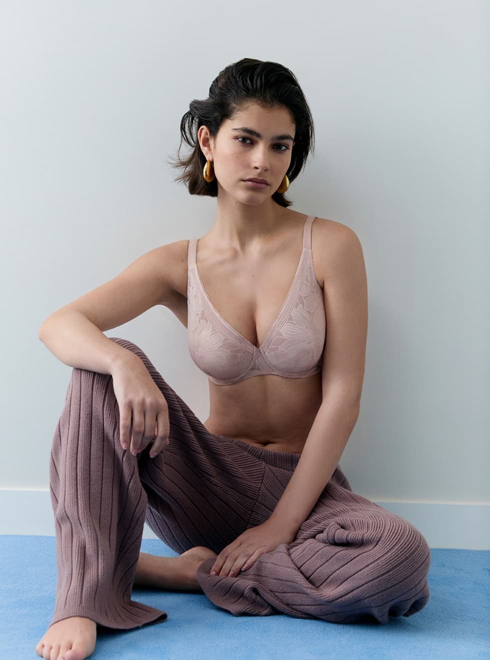 Woman sitting on blue rug, wearing a light bra and brown knitted pants, with gold hoop earrings against a white wall.