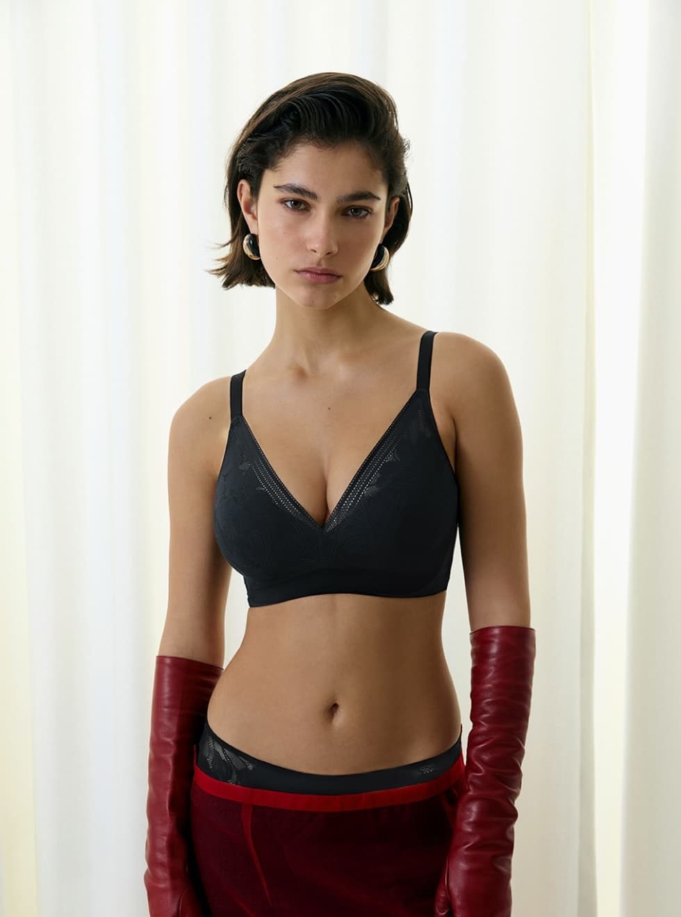 Woman in a black bra and red elbow-length gloves stands against a light curtain background.