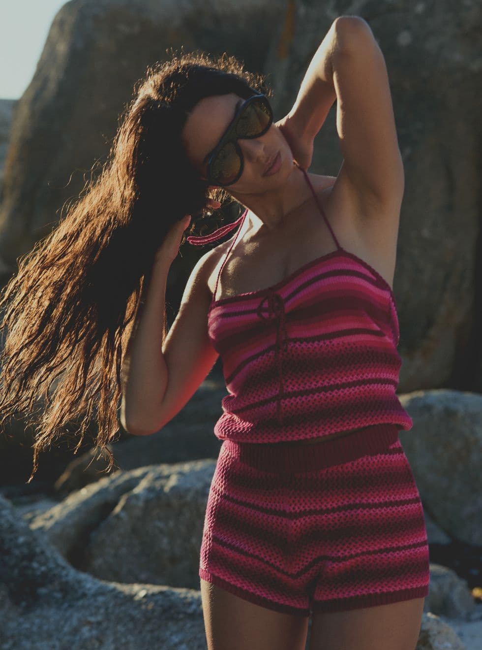 Woman in a pink-striped outfit and sunglasses, touching her hair, poses outdoors by rocks in sunlight.