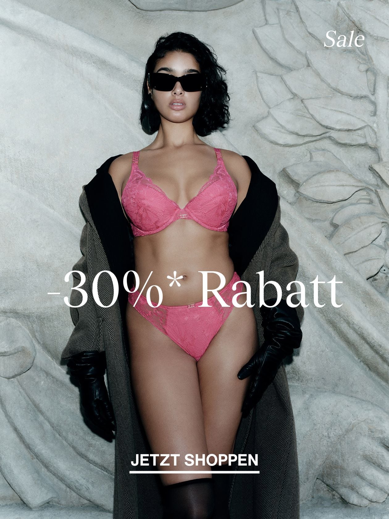 Model in pink lace bra and panties, oversized coat and black gloves, wearing sunglasses, posing against carved stone backdrop with sale text.