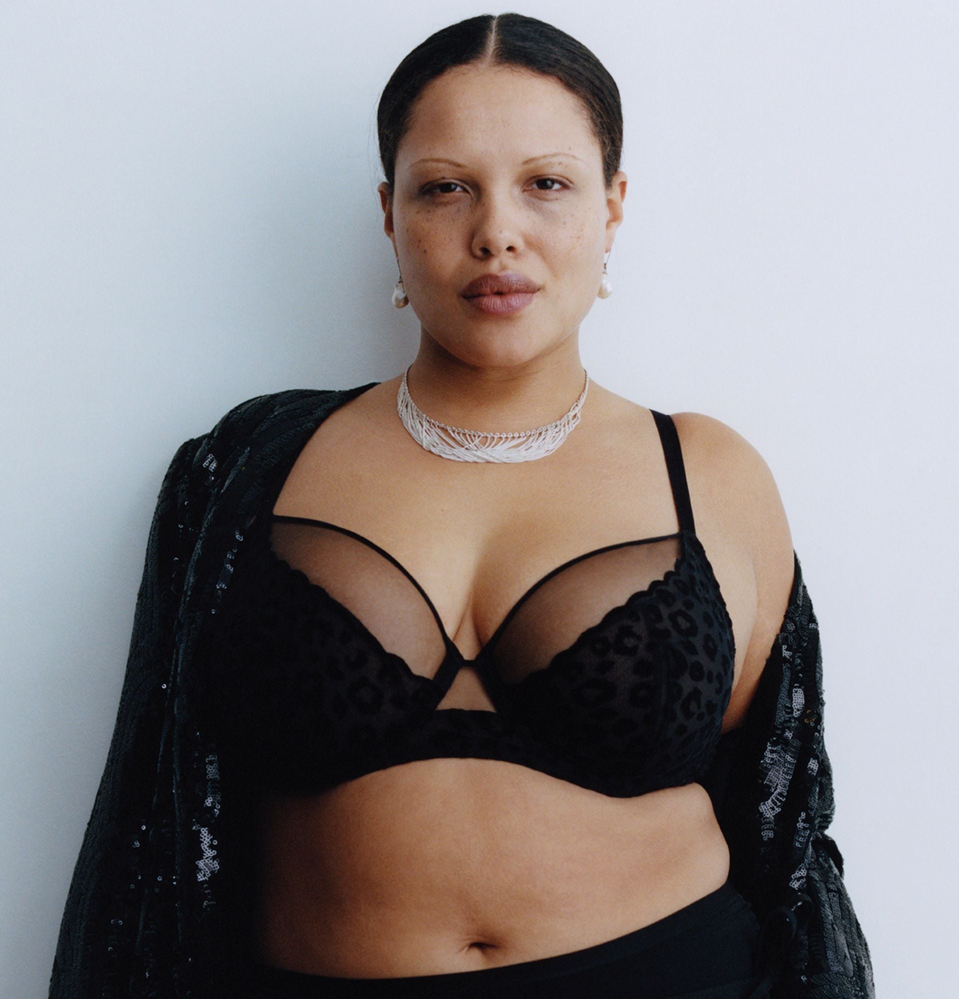A woman is shown wearing a black sheer bra with a subtle animal print, layered jewelry, and a draped black garment, standing confidently against a plain backdrop.