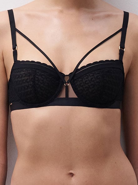Person wearing a black lace bra with decorative straps and scalloped edges, standing against a plain background.