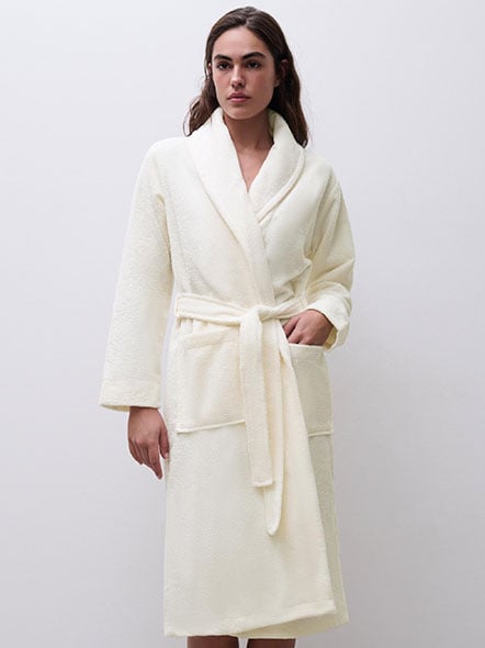 Person wearing a cream-colored bathrobe with a shawl collar and belt, standing against a plain white background.