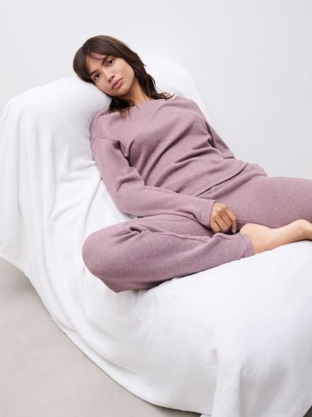Person in a mauve loungewear set sits relaxed on a white-covered chair, with a neutral expression against a plain background.