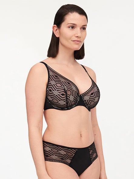 A woman models a black bra and panties set with geometric lace patterns, standing against a plain white background.