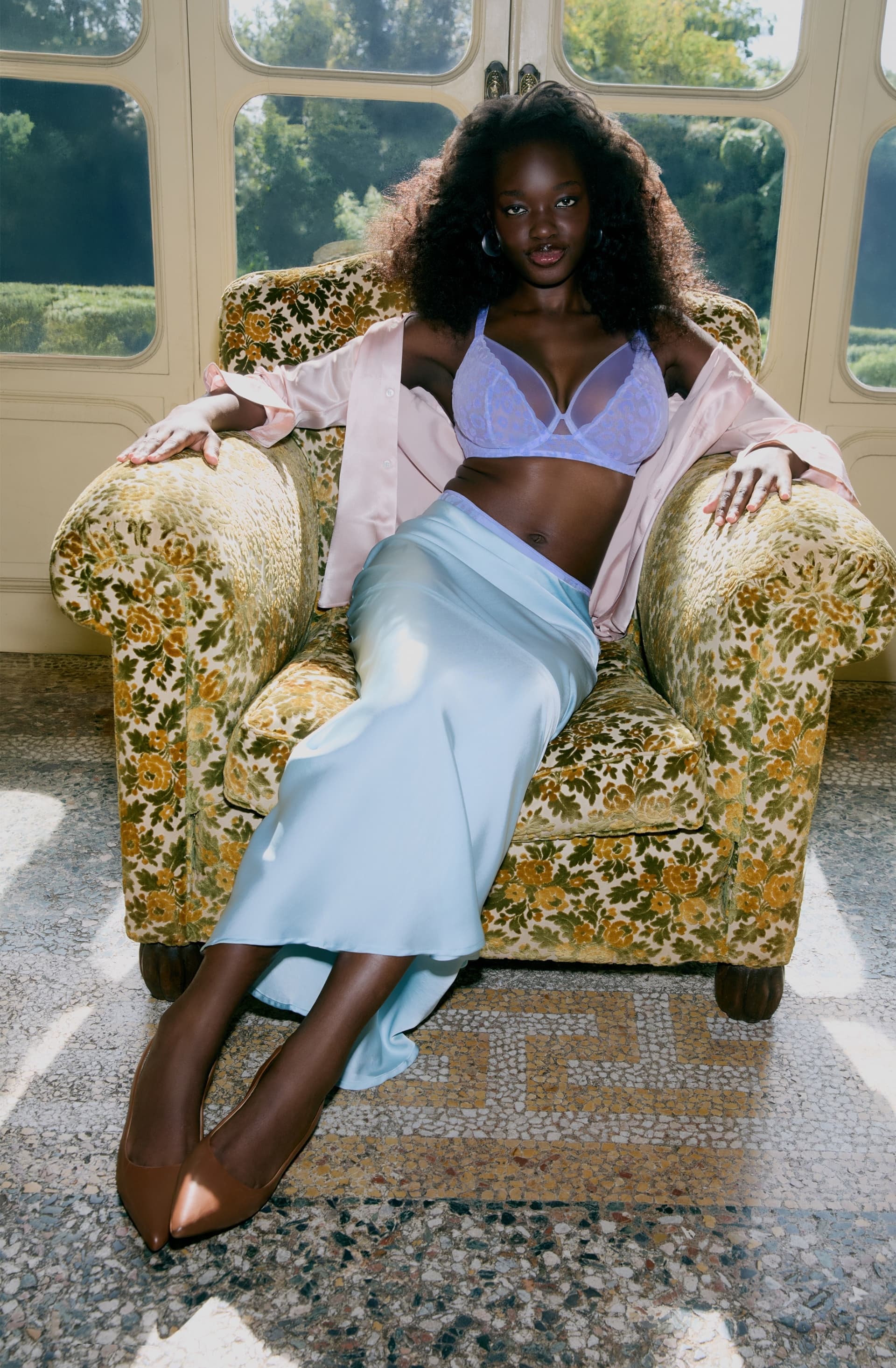 A woman in a lilac bra, satin outfit, and brown heels sits on a floral armchair in a sunlit room with large windows and a garden view.