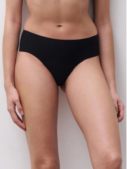 Person wearing high-waisted black underwear, standing against a light wall, showcasing a minimalistic garment style.