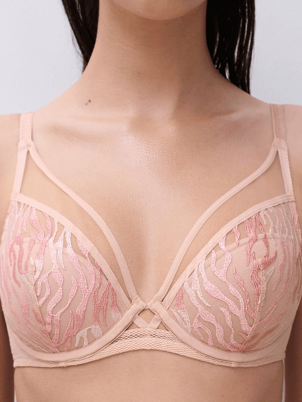 A close-up of a woman wearing a pink and beige lace bra with delicate embroidery and thin shoulder straps.