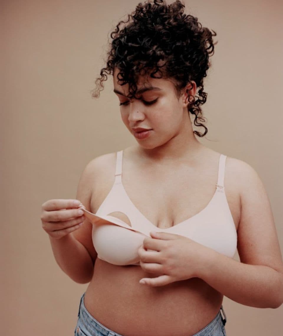 Woman with curly hair wearing a nude bra and jeans, standing against a plain beige background.