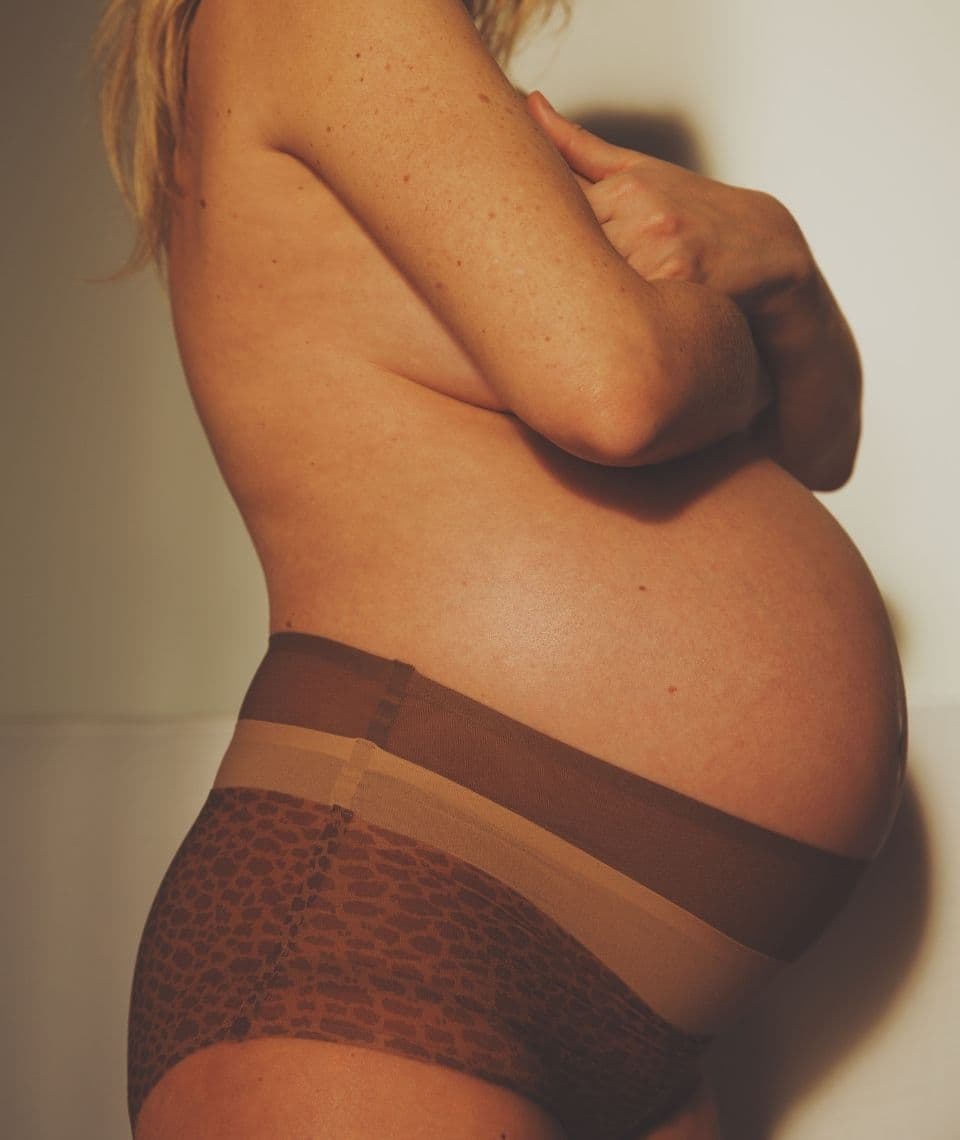 Pregnant person in animal print underwear, torso visible, with hands crossed over chest, standing against a light background.