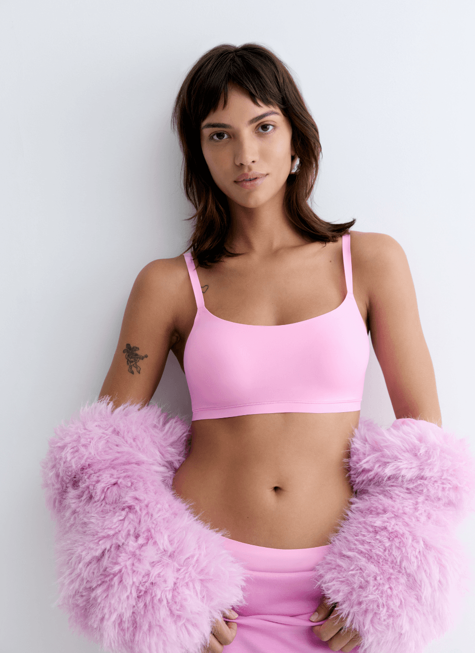 A person wearing a pink bra and skirt, draped in pink fur, standing against a white background, with a visible arm tattoo.
