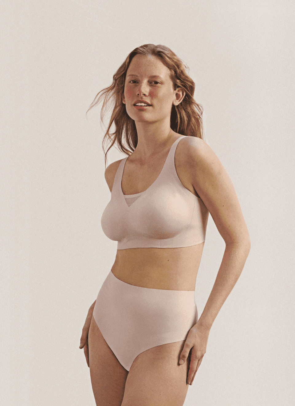 A person in nude-colored bra and high-waisted underwear stands against a plain background, looking confidently at the camera.