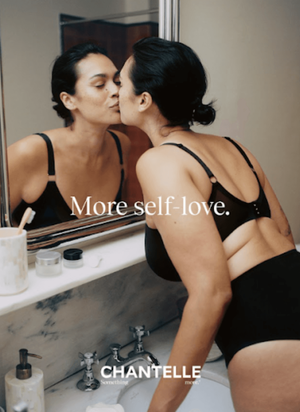 A woman in black underwear kisses her reflection in a bathroom mirror with the words "More self-love" overlayed. Products are on the counter.