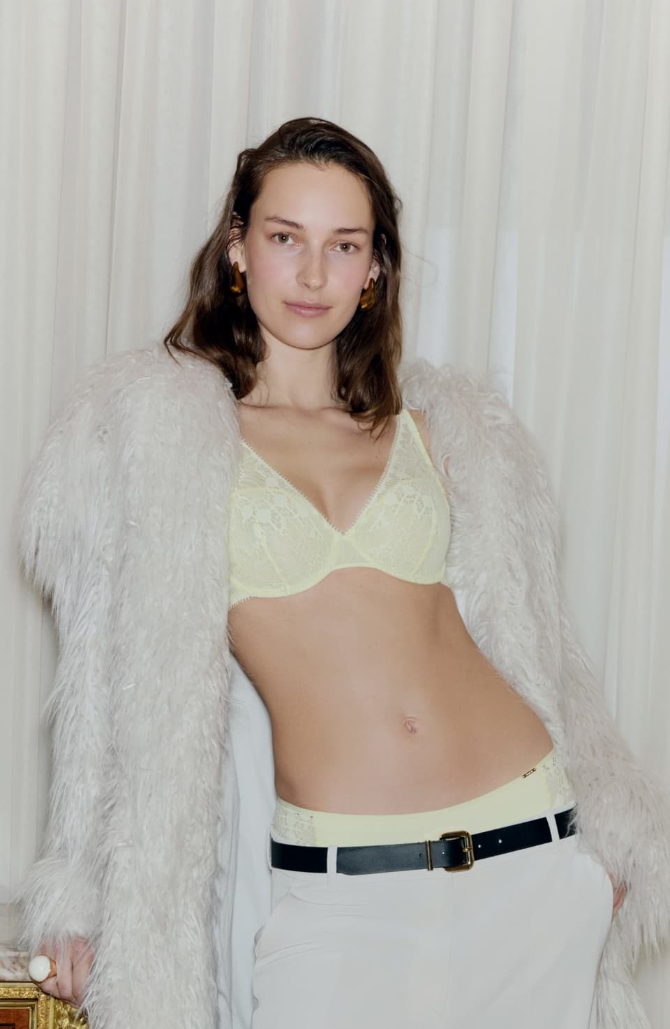 Person wearing a pale yellow lace bra, cream pants, and a fluffy coat, standing by white curtains with one hand on their hip.