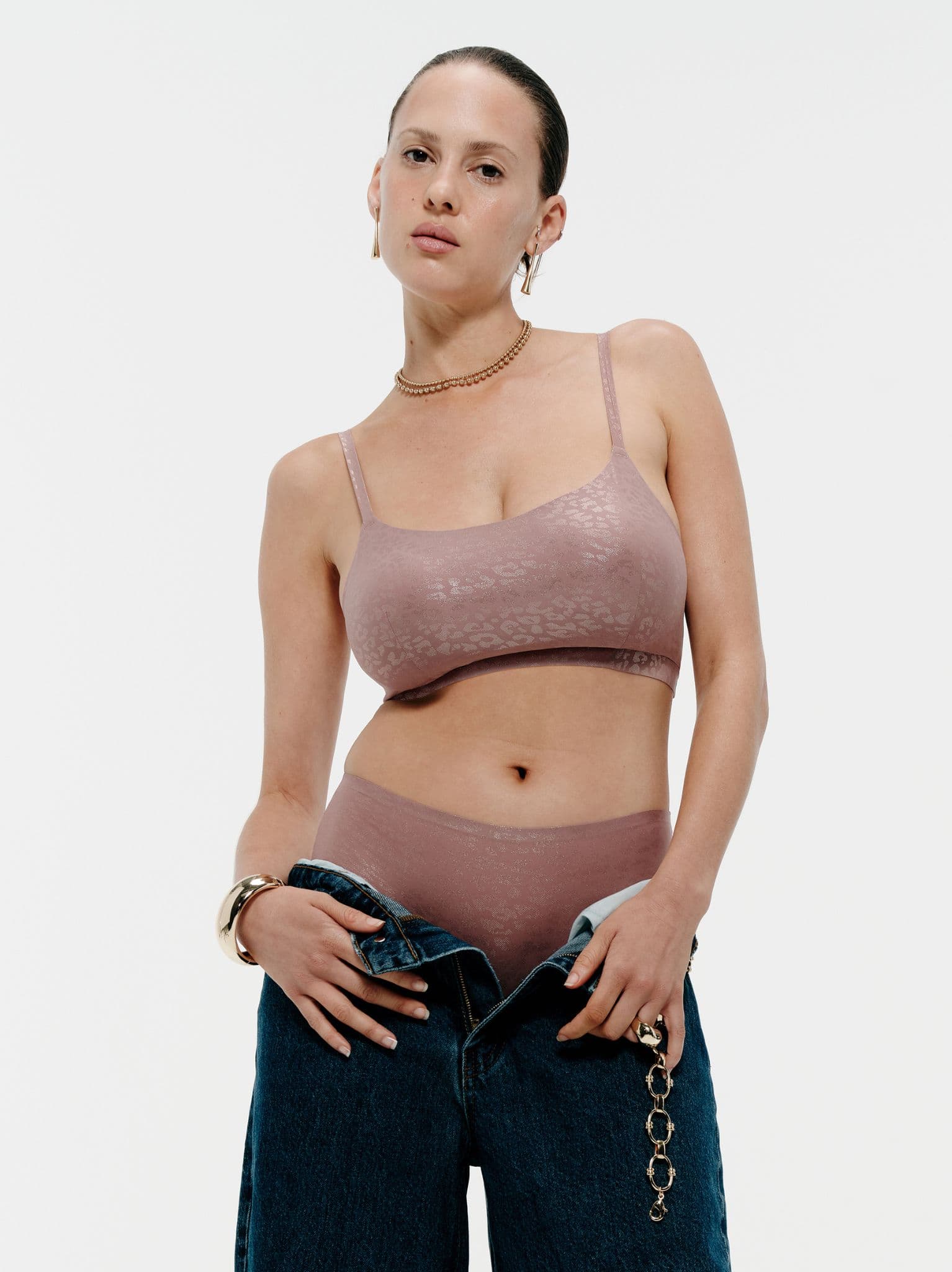 Person wearing a mauve bralette and underwear, holding a pair of unbuttoned jeans. They have short hair and wear gold jewelry.