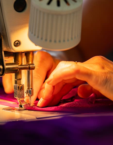 Mains cousant du tissu violet à l'aide d'une machine à coudre, en mettant l'accent sur les détails de l'aiguille et des coutures sous un éclairage chaleureux.