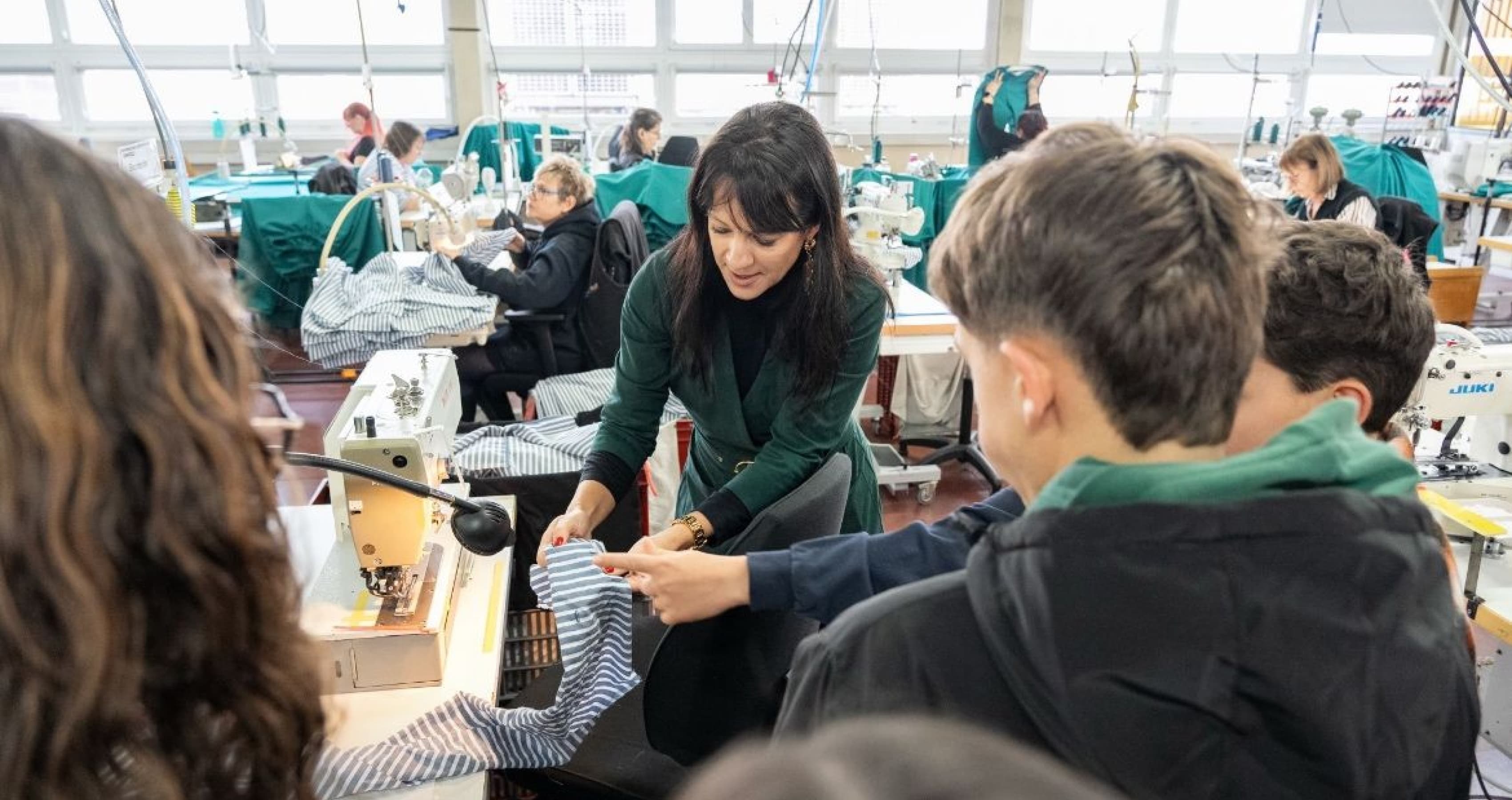 Un groupe de personnes observe la responsable de l'atelier de confection qui fait une démonstration de techniques de couture dans un atelier textile. On aperçoit des machines à coudre et des tissus.